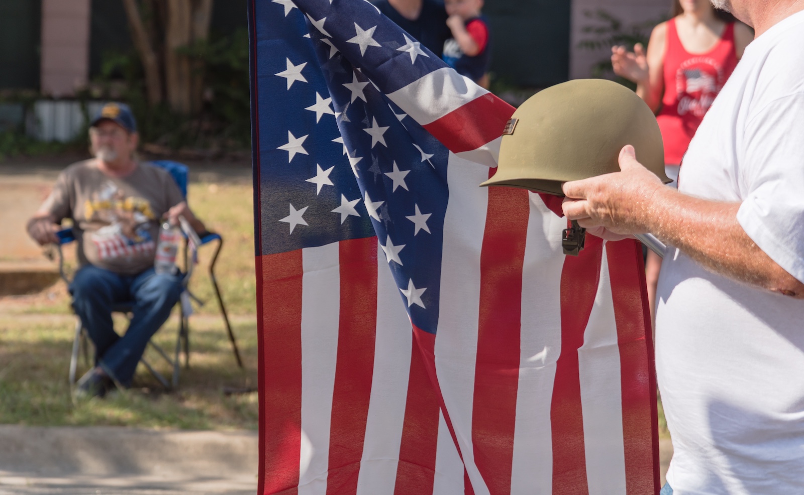 Side view a white veteran proudly holding military WWI helmet (M1 helmet) and US flag. July 4th or veterans day poster of WWII, modern wars. American soldier troop during parade with people watching